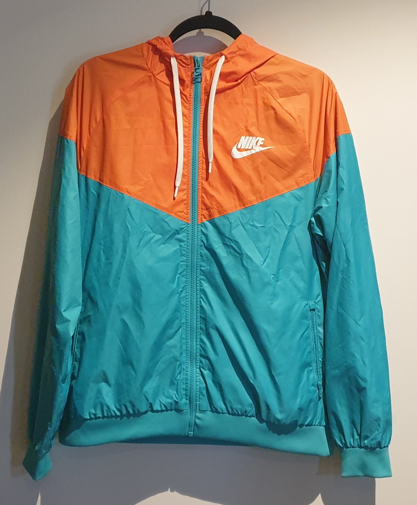 A Nike sprayer jacket with long sleeves, featuring orange color on the upper part and blue color on the lower part. The jacket has a white Nike logo on the chest and is displayed on a hanger against a plain background.